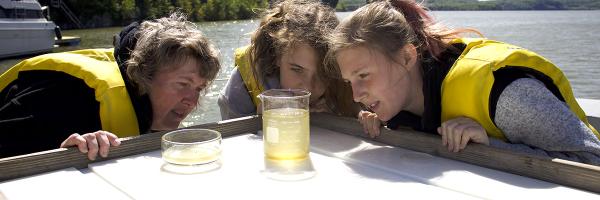 A teacher and her students examine a phytoplankton sample aboard the University of Vermont research vessel on Lake Champlain. Photo: Ashley Eaton A teacher and two students in yellow life vests examine a water sample in a beaker.